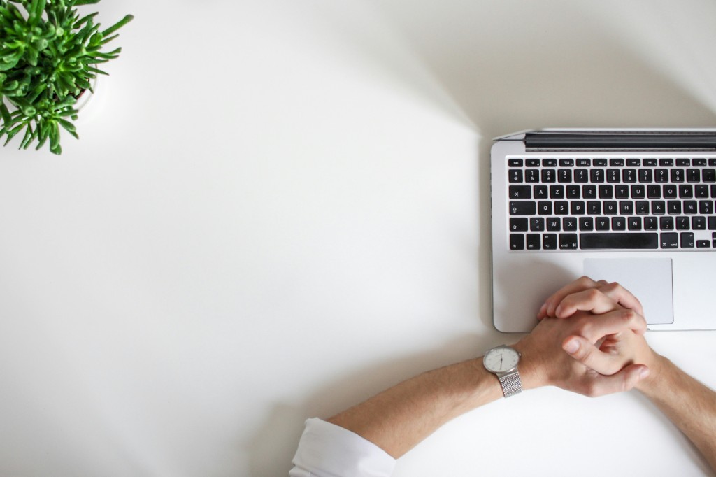 Overhead view of hands resting beside a laptop on a white desk